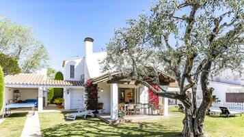 A white villa with a tiled roof, a covered patio with outdoor furniture, and a large tree in the foreground.