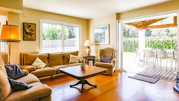 A living room with a sofa, coffee table, and armchairs. There is a window with a view of greenery outside.