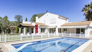 A swimming pool with a white fence, a white building with a tiled roof, and a palm tree in the background. - Casa Independiente con Jard n y Piscina Privada (Sant Pere de Ribes)