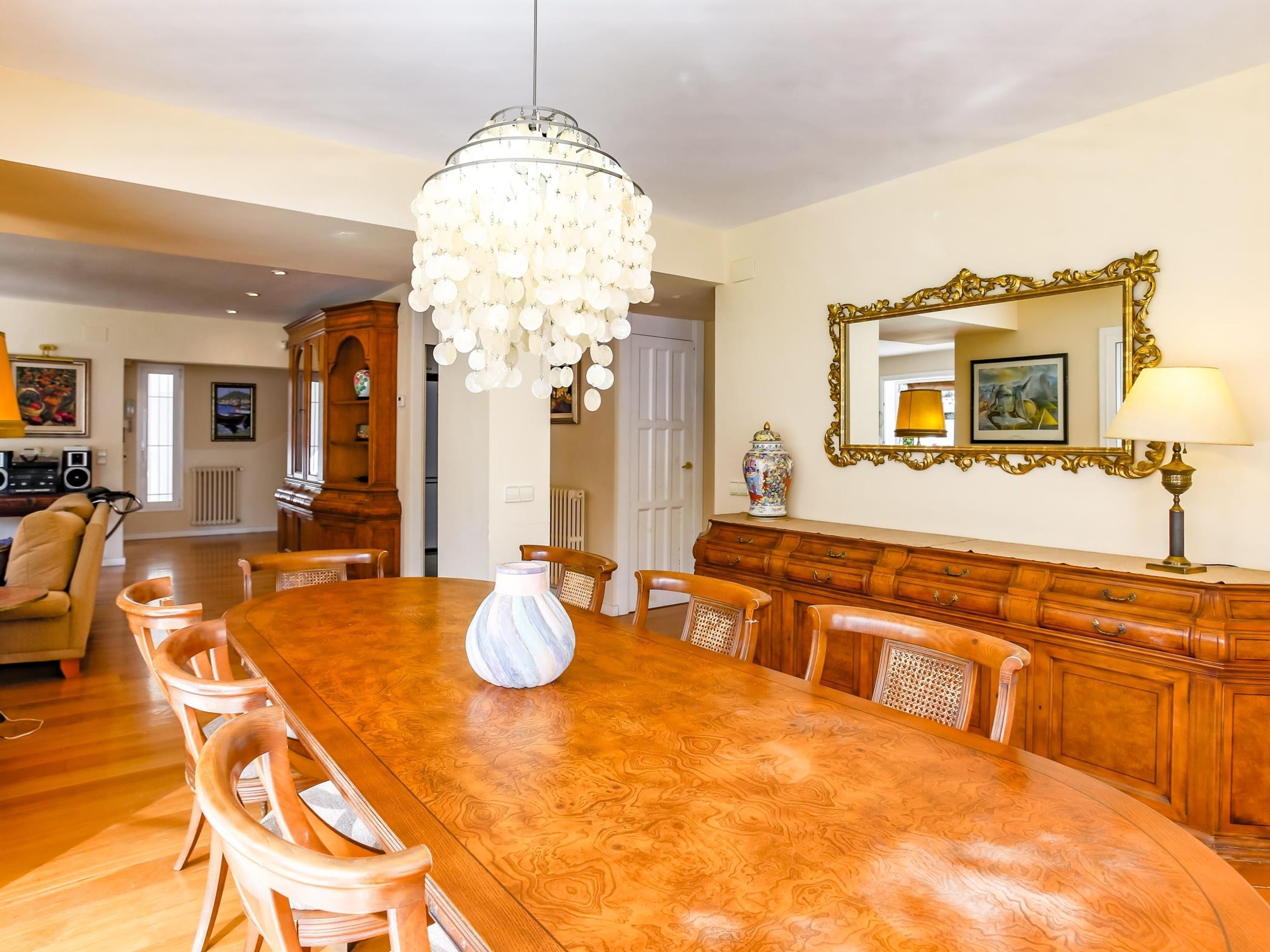 A dining room with a large wooden table, chairs, a chandelier, a mirror, and a cabinet.