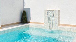 Una piscina con caída de agua, un árbol pequeño y una lámpara fijada a la pared.