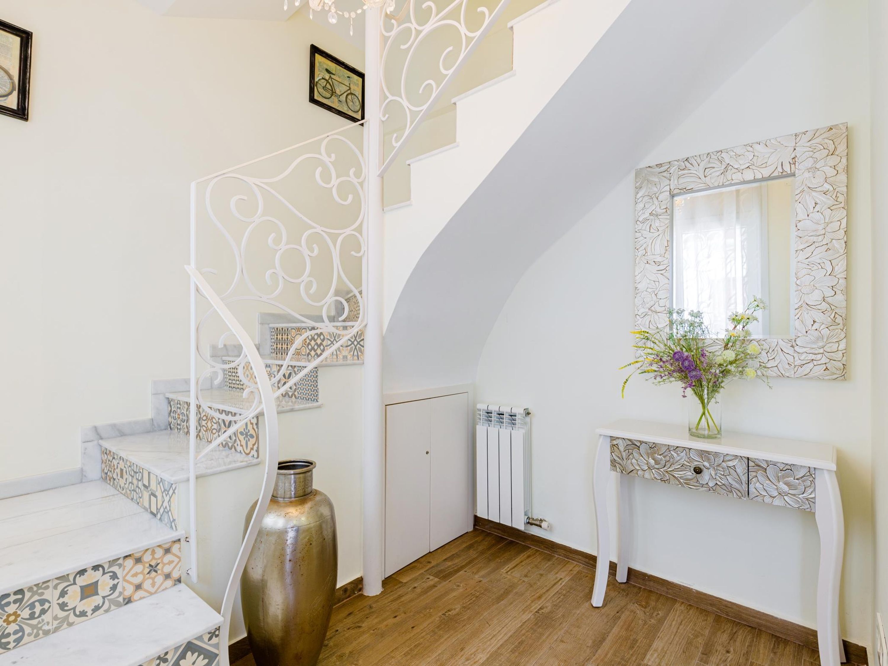 A staircase with white railings and patterned steps, a radiator, a console table with a mirror, and a vase with flowers.