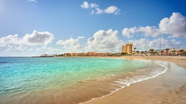 A beach with clear turquoise water, a sandy shore, and a row of buildings in the background. - C Ntrico y a pie de Playa en los Cristianos (Arona)