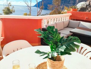 Ένα μπαλκόνι με ένα λευκό τραπέζι, δύο ποτήρια, ένα φυτό σε γλάστρα και έναν καναπέ.