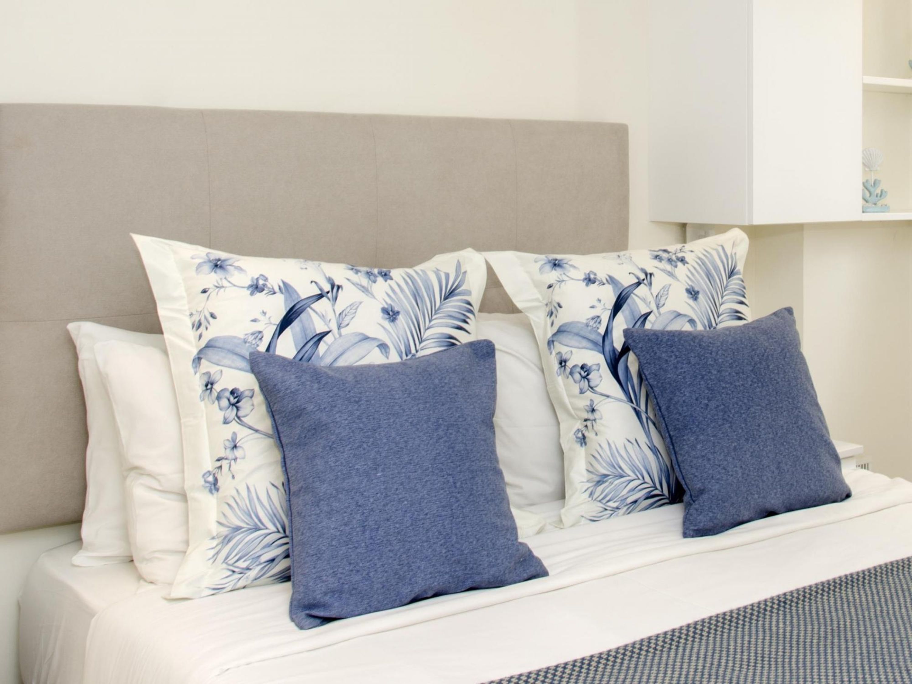 Una cama con cabecera color beige, ropa de cama blanca y dos almohadas con estampado floral azul.