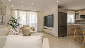 A modern living room with a sofa, a coffee table, and a TV. There is a kitchen area with a refrigerator, microwave, and sink. - Sitges Spaces Oasis Suites (Sitges)