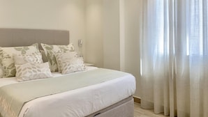 A bedroom with a bed, pillows, a headboard, a window with curtains, and a wall-mounted light. - Sitges Spaces Oasis Suites (Sitges)