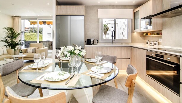 A modern kitchen with a central island, a dining area with a glass table, and a view of the outdoors. - Sitges Spaces Oasis Suites (Sitges)