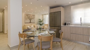 A modern dining area with a glass table surrounded by wooden chairs, a large refrigerator, and a kitchen counter with a sink. - Sitges Spaces Oasis Suites (Sitges)