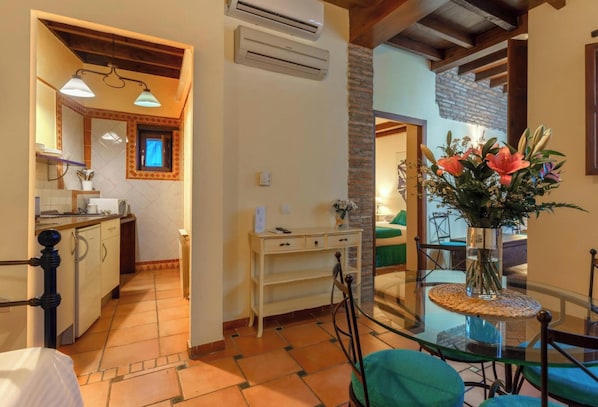A cozy room with a bed, a glass table with chairs, a vase of flowers, and a view of a kitchen and dining area.