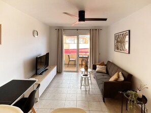 A living room with a TV, a sofa, a dining table, and a ceiling fan. - 21 Relax Oasis del mar (Santiago del Teide)