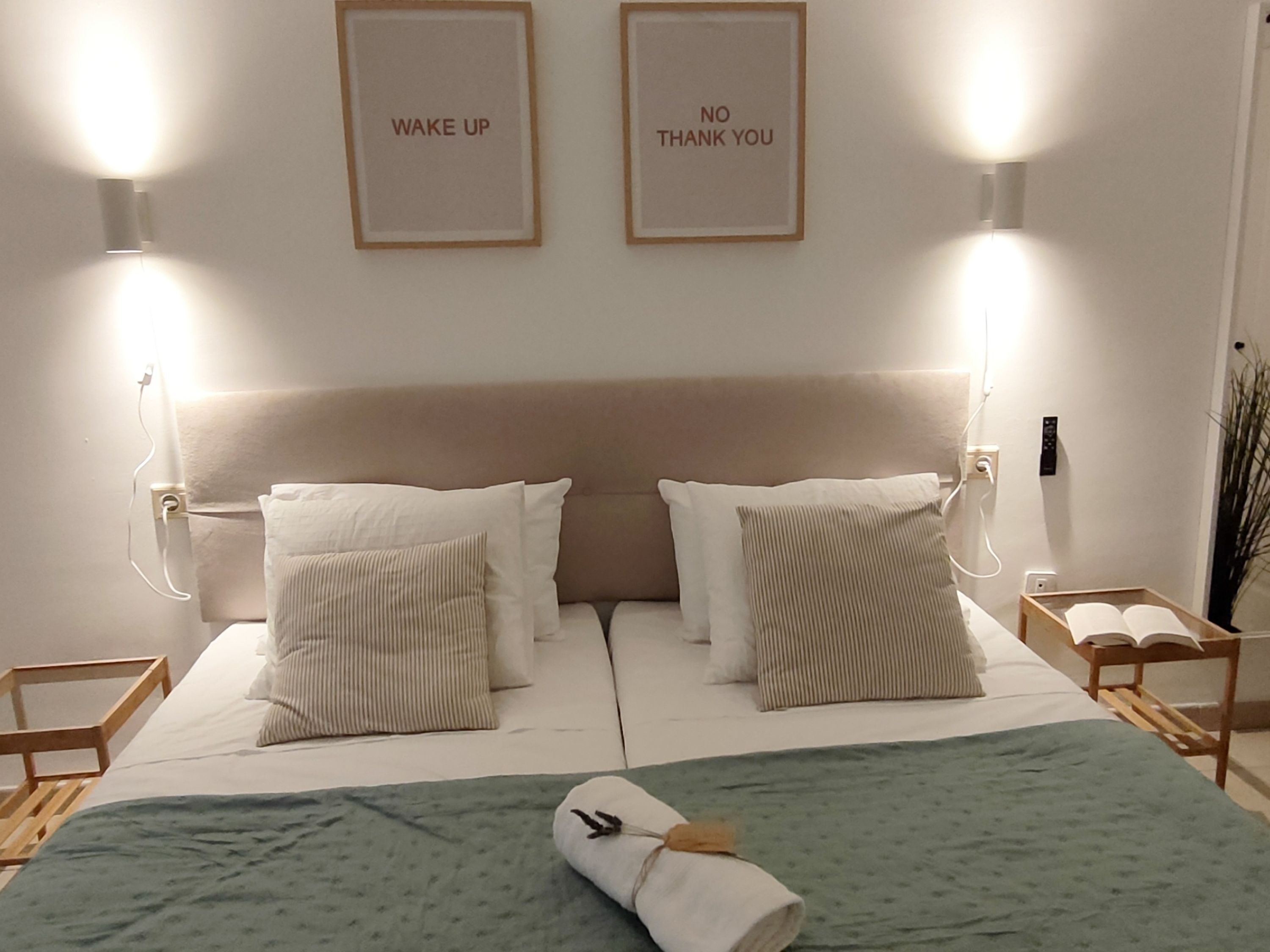 Ein ordentlich bezogenes Bett mit Kissen, einer grünen Decke und einem zusammengerollten Handtuch. Es befinden sich zwei Wandleuchten mit Beschriftung an der Wand.