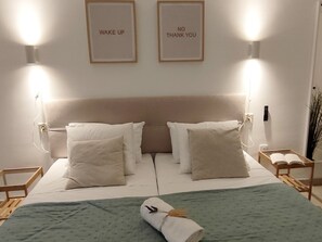 Un lit bien fait, avec des oreillers, une couverture verte et une serviette enroulée. Deux appliques murales avec des inscriptions y sont fixées.