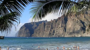 A beach with people swimming and standing on the sand, palm trees, and a rocky cliff in the background.