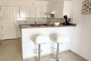 A kitchen with white cabinets, a granite countertop, two white bar stools, and a bowl of fruit.