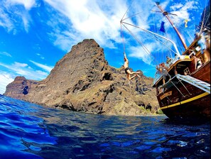 Un navire avec une personne qui fait du saut à l’élastique depuis la proue, une île rocheuse en arrière-plan et une eau d’un bleu limpide.