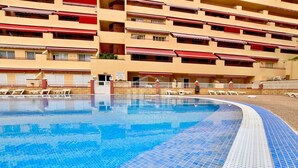 A swimming pool with a tiled edge, surrounded by lounge chairs and a building with balconies and awnings.