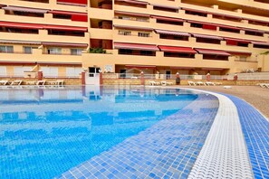 Une piscine dont le bord est revêtu de carreaux, entourée de chaises longues et bordée d’un bâtiment doté de balcons et d’auvents.