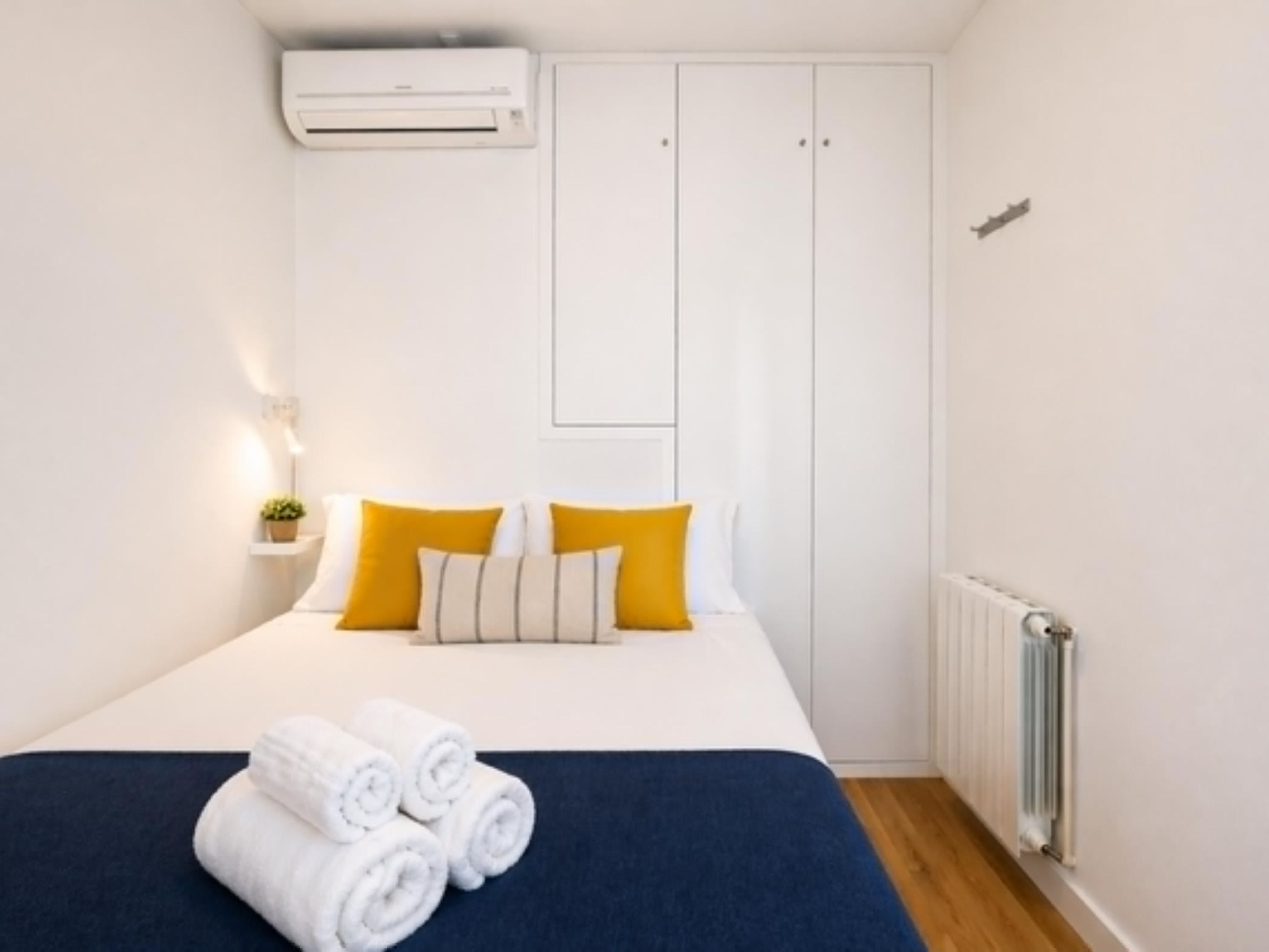 Un dormitorio con cama, paredes blancas, un equipo de aire acondicionado, un radiador y una planta pequeña en una mesita de noche.