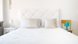 Een net opgemaakt bed met een gewatteerd hoofdbord, een bedlampje en een klein tafeltje met een lamp.