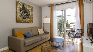 A living room with a sofa, a round table, and two chairs. There is a painting on the wall, a lamp, and a window with curtains. - Sitges Spaces Terrace Apartment (Sitges)