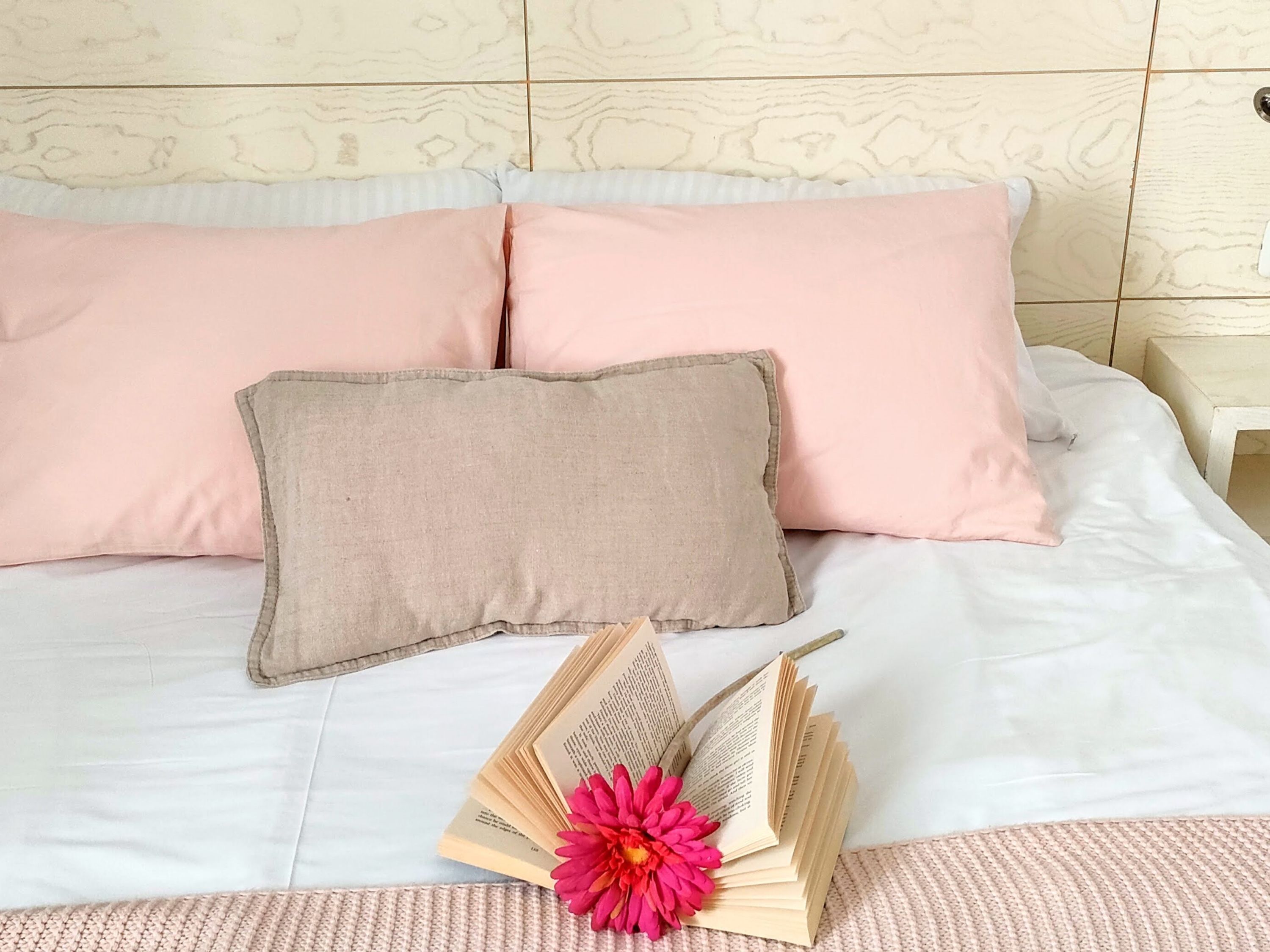 A bed with pink and beige pillows, a knitted throw, and a book with a pink flower on it.