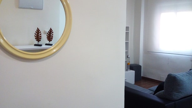 A living room with a round mirror, a sofa, a small table with decorative items, and a window with blinds. - Luminous Flat in Great Location (Valencia)