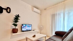 Ett vardagsrum med en soffa, en tv som står på en hylla, en växt och en fläkt.