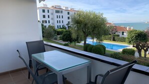A balcony with a glass table and chairs, overlooking a swimming pool and a residential area with white buildings and trees. - Ground Floor in Sitges (Sitges)