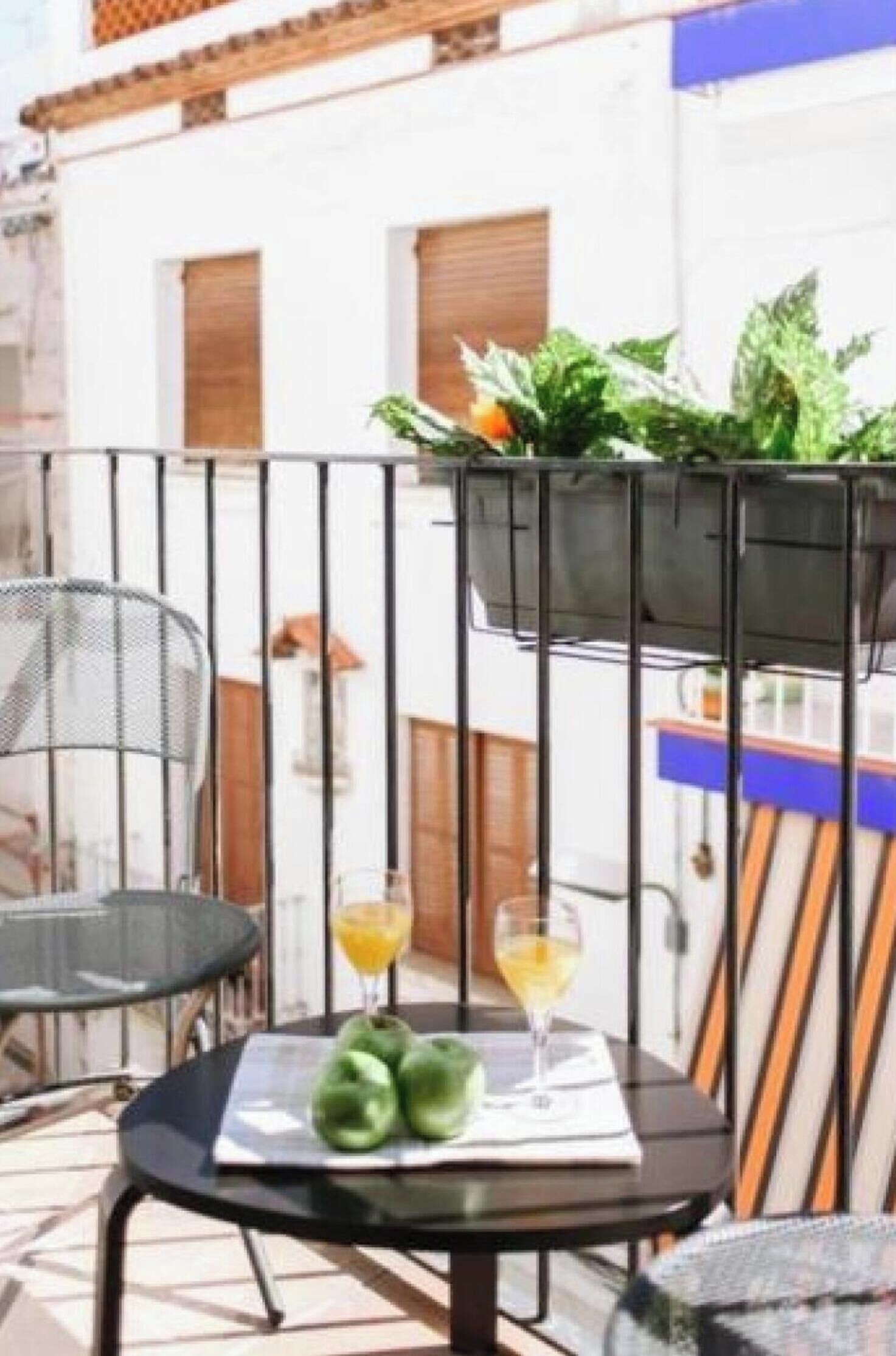 Un balcón con una mesa, dos sillas, una silla metálica y un macramé colgante con plantas verdes.