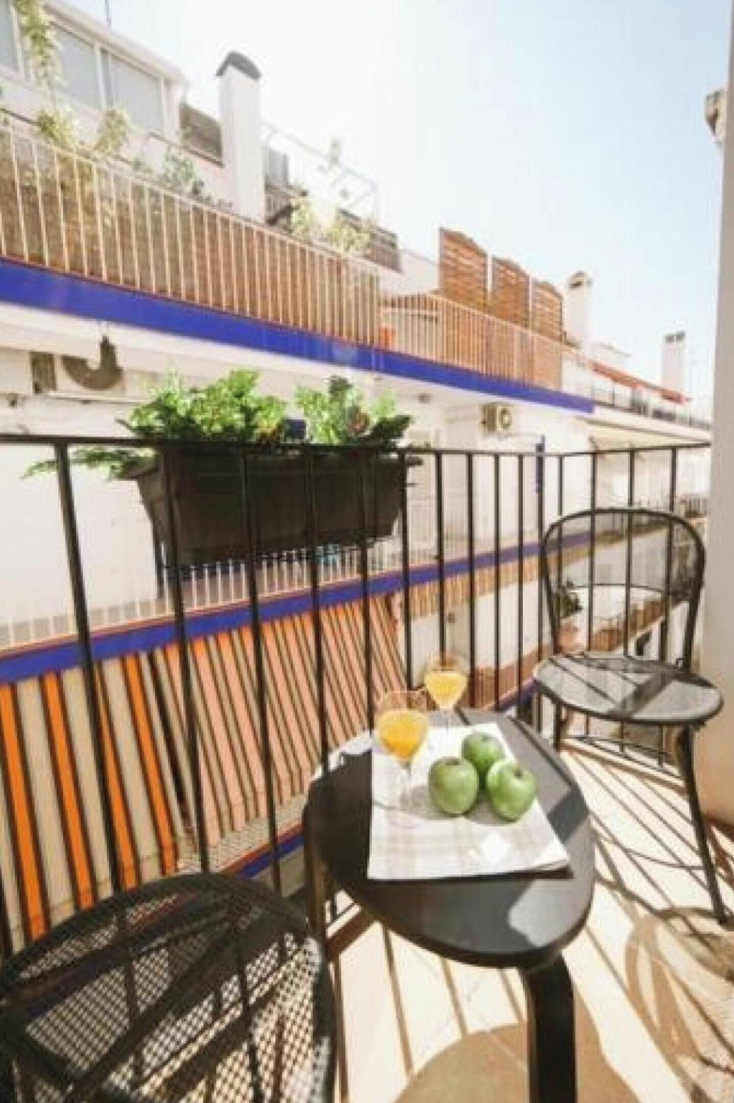 Un balcón con una mesa dispuesta con frutas y una bebida, una silla y una maceta colgante con plantas.