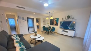 A modern living room with a sofa, a dining table with chairs, a TV, and a large painting on the wall.