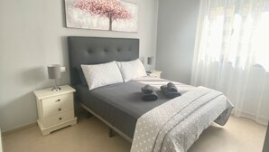A modern bedroom with a grey upholstered bed, a bedside table with a lamp, a small white dresser, and a large window with sheer curtains.