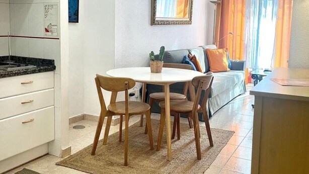 A compact living space with a kitchenette, a dining area with a round table and chairs, a sofa with cushions, and a large window with curtains. - C Ntrico y Acogedor Ideal Para Explorar la Ciudad (Alicante)