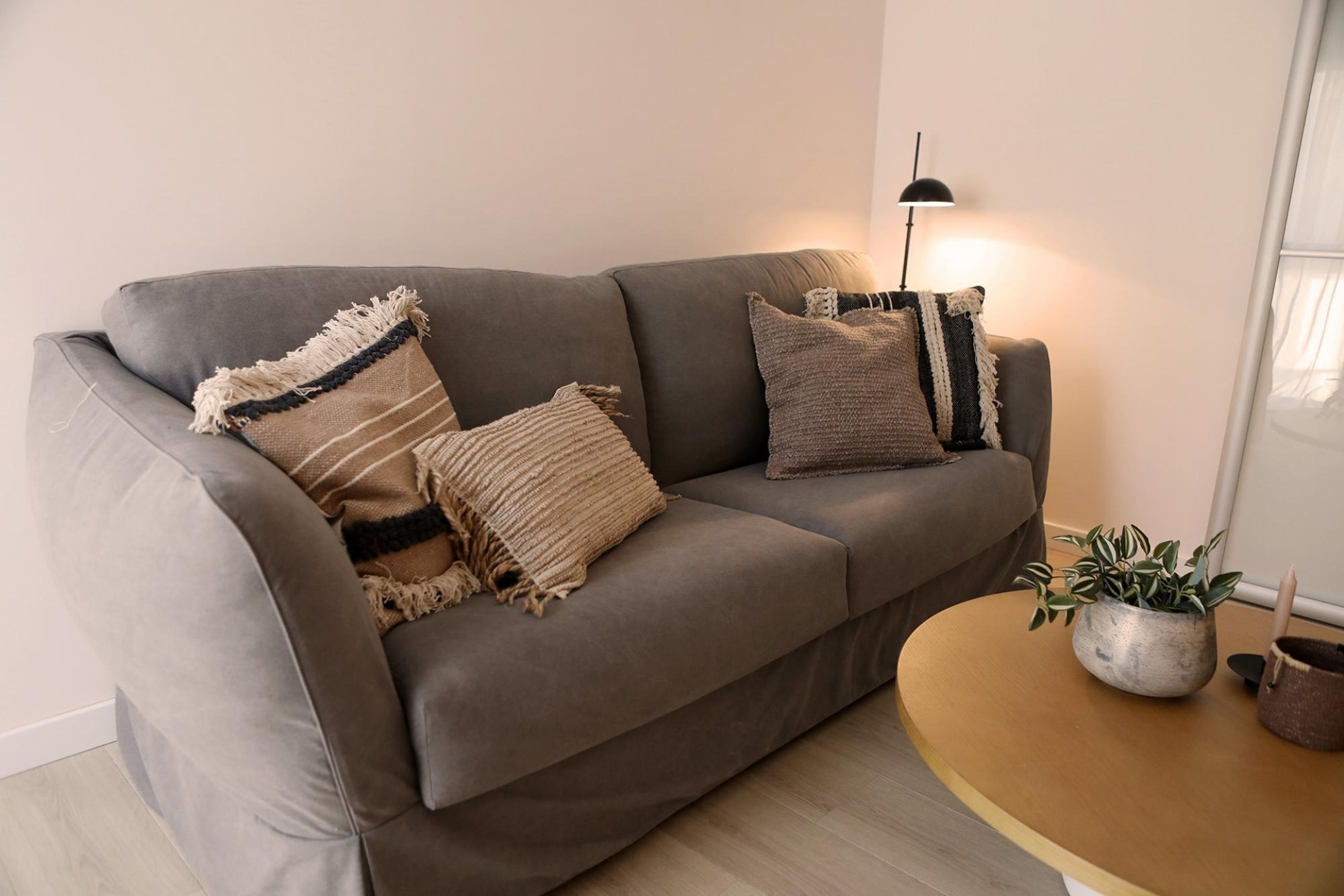 A grey sofa with three cushions, a small round wooden table with two potted plants, and a floor lamp.