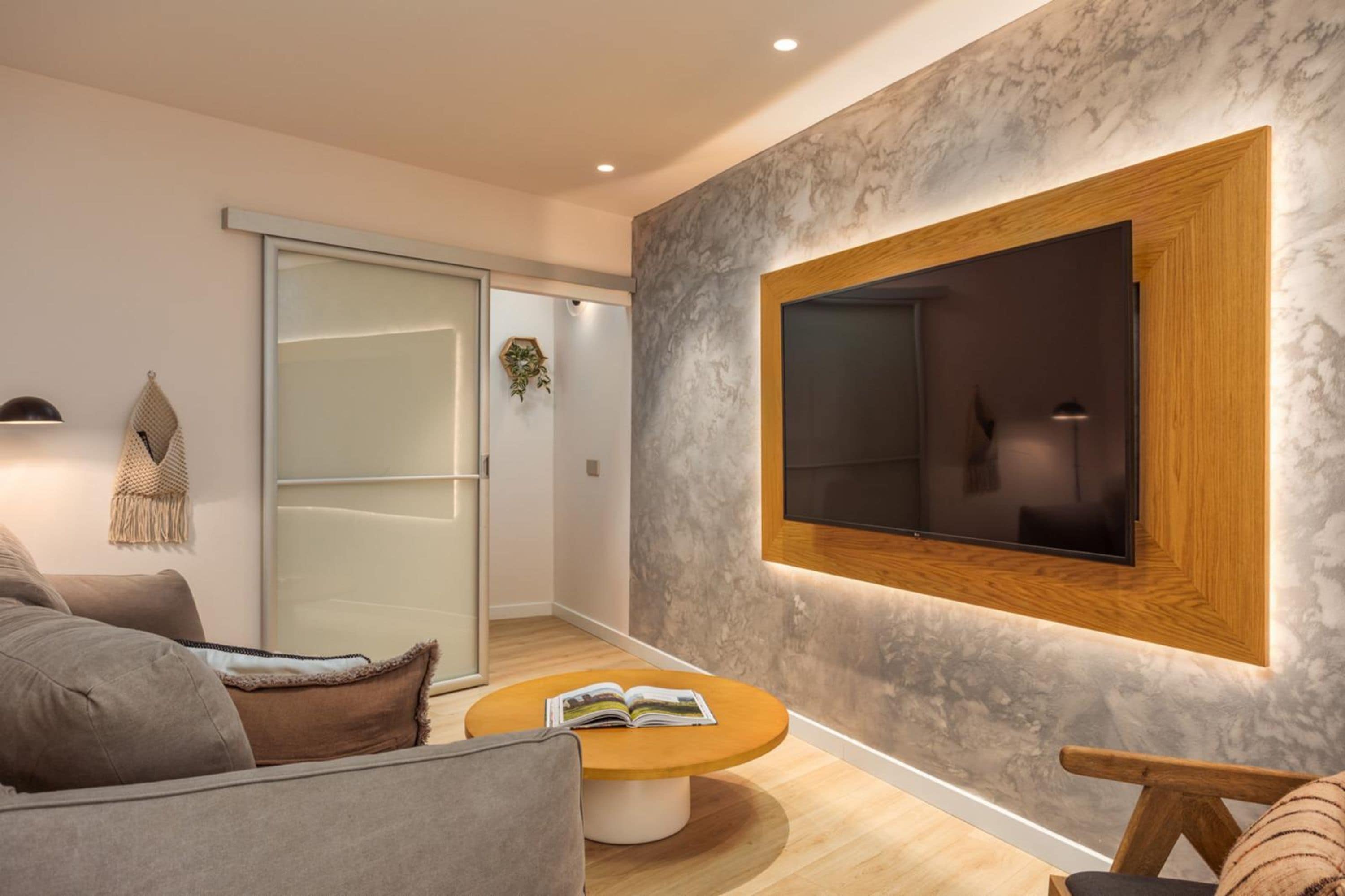 A modern living room with a large flat-screen TV mounted on a textured wall, a sliding glass door leading to another room, and a round wooden coffee table with a book.