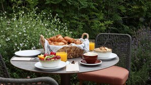 An outdoor dining table with croissants, fruit, juice, and coffee, surrounded by greenery.