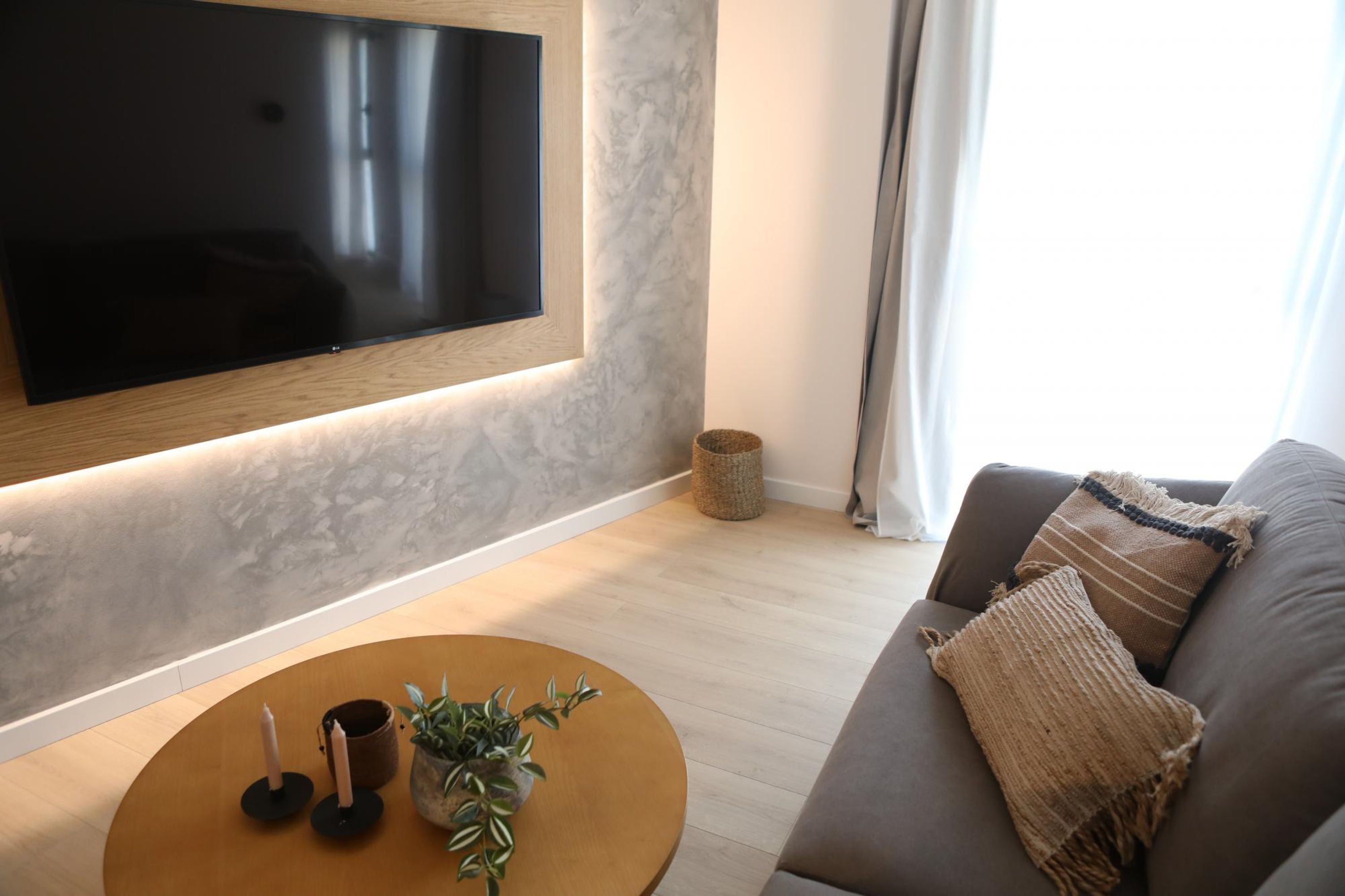 A modern living room with a flat-screen TV mounted on the wall, a round wooden coffee table with candles and a plant, a grey sofa with a textured throw blanket, and a wicker basket on the floor.