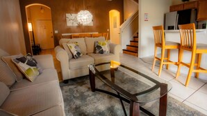 A living room with a sofa, a glass coffee table, and a kitchen area with wooden chairs and cabinets. - Where Memories Flourish 8 Mins to Disney (Davenport)