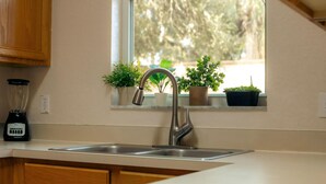 A kitchen with a stainless steel sink, wooden cabinets, a window with potted plants, and a blender on the counter. - Where Memories Flourish 8 Mins to Disney (Davenport)