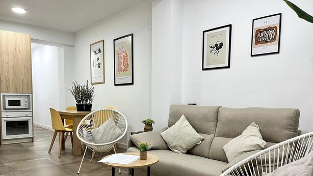 A modern living room with a sofa, two white wicker chairs, a small table, and a dining area with a chair and a plant.