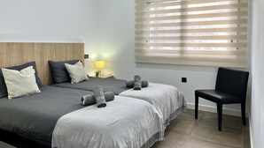 A modern bedroom with two beds, a nightstand with a lamp, a chair, and a large window with blinds.