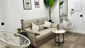 A modern living room with a beige sofa, two white wicker chairs, a small wooden side table, and a large green plant.