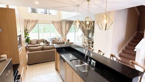 A modern kitchen with a black countertop, wooden cabinets, and a built-in sink. There is a staircase leading to an upper level, and a dining area with a sofa and chairs.