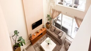 A modern living room with a flat-screen TV, a sofa, a coffee table, and a large window offering a view of the outdoors. - Experience the Charm of Snowman 5 Miles to Disney (Kissimmee)