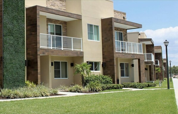 A modern multi-story residential building with balconies, a well-maintained lawn, and a clear sky.