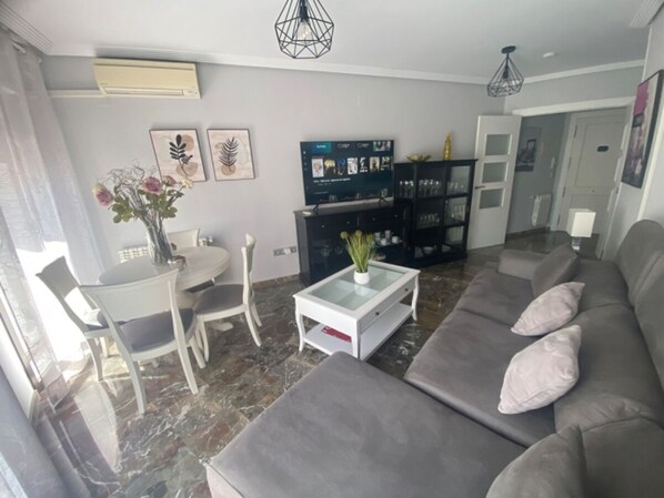 A modern living room with a grey sofa, a coffee table, and a dining area with a white table and chairs. - Bonito Apartamento en Centro Ciudad con Garaje (Albaida)