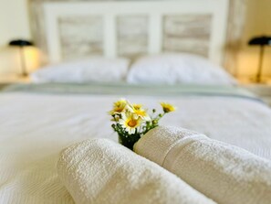 A neatly made bed with a white comforter and pillows, a small bouquet of yellow flowers, and a headboard with a light wood finish.