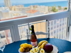A table with a bottle of cava, lemons, bananas, a peach, and daisies on a balcony with a city view.