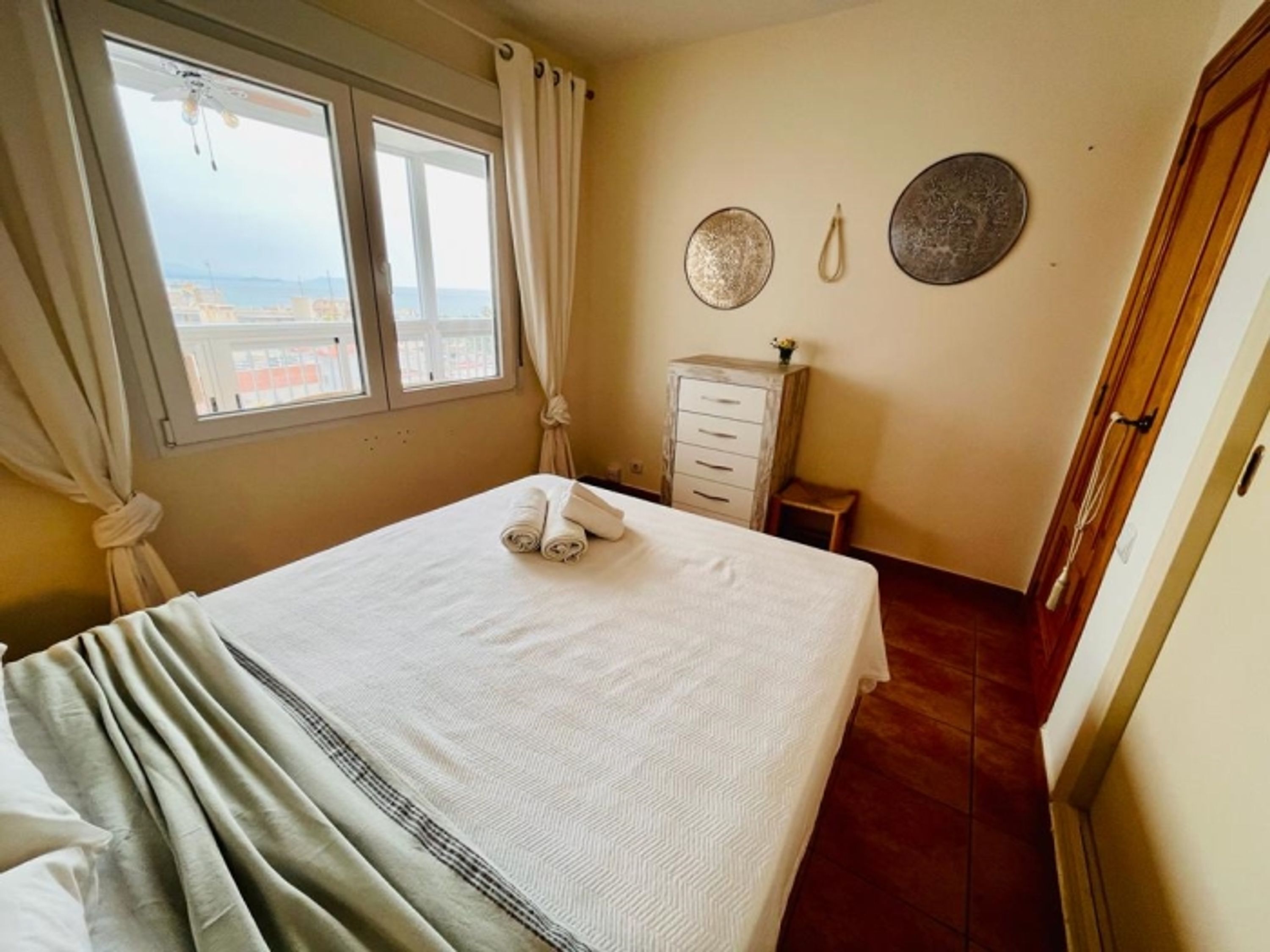 A neatly made bed with a white bedspread, a window with curtains, a wooden dresser, and a wall with two decorative plates.
